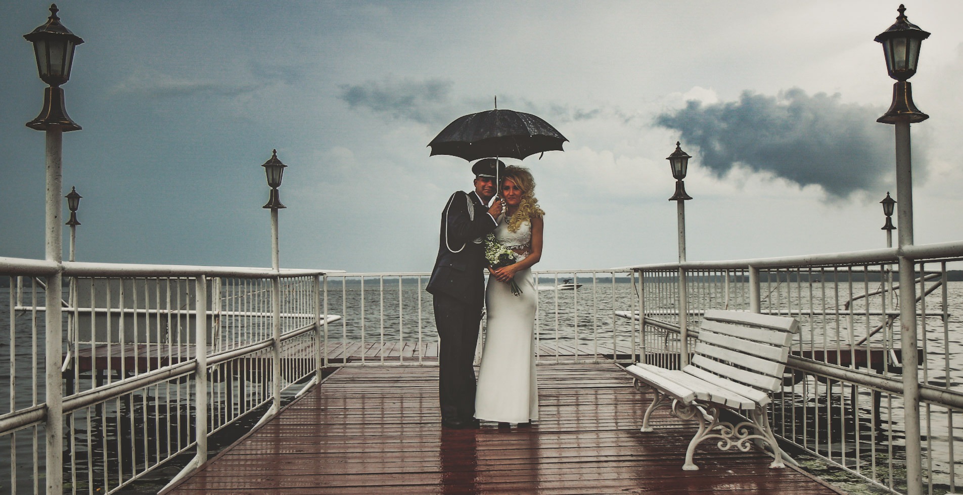 Para młoda w deszczu na molo, pan młody w mundurze trzyma parasol, w tle pochmurne niebo i jezioro.