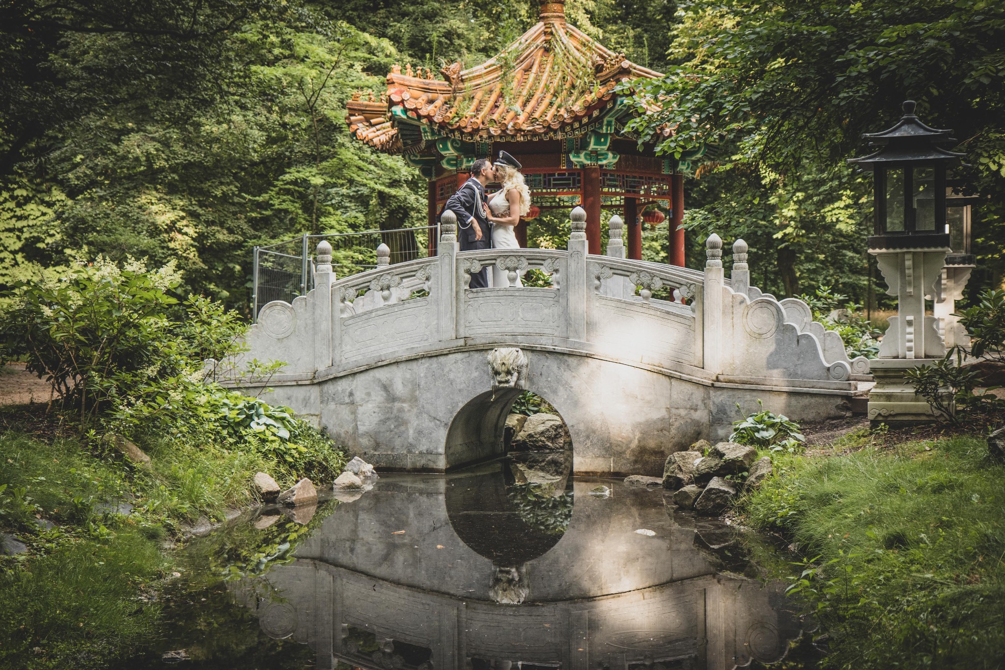 Para młoda w nietypowych strojach (on w mundurze, ona w białej sukni) całuje się na kamiennym moście w orientalnym ogrodzie z pagodą w tle, odbicie mostu w wodzie.