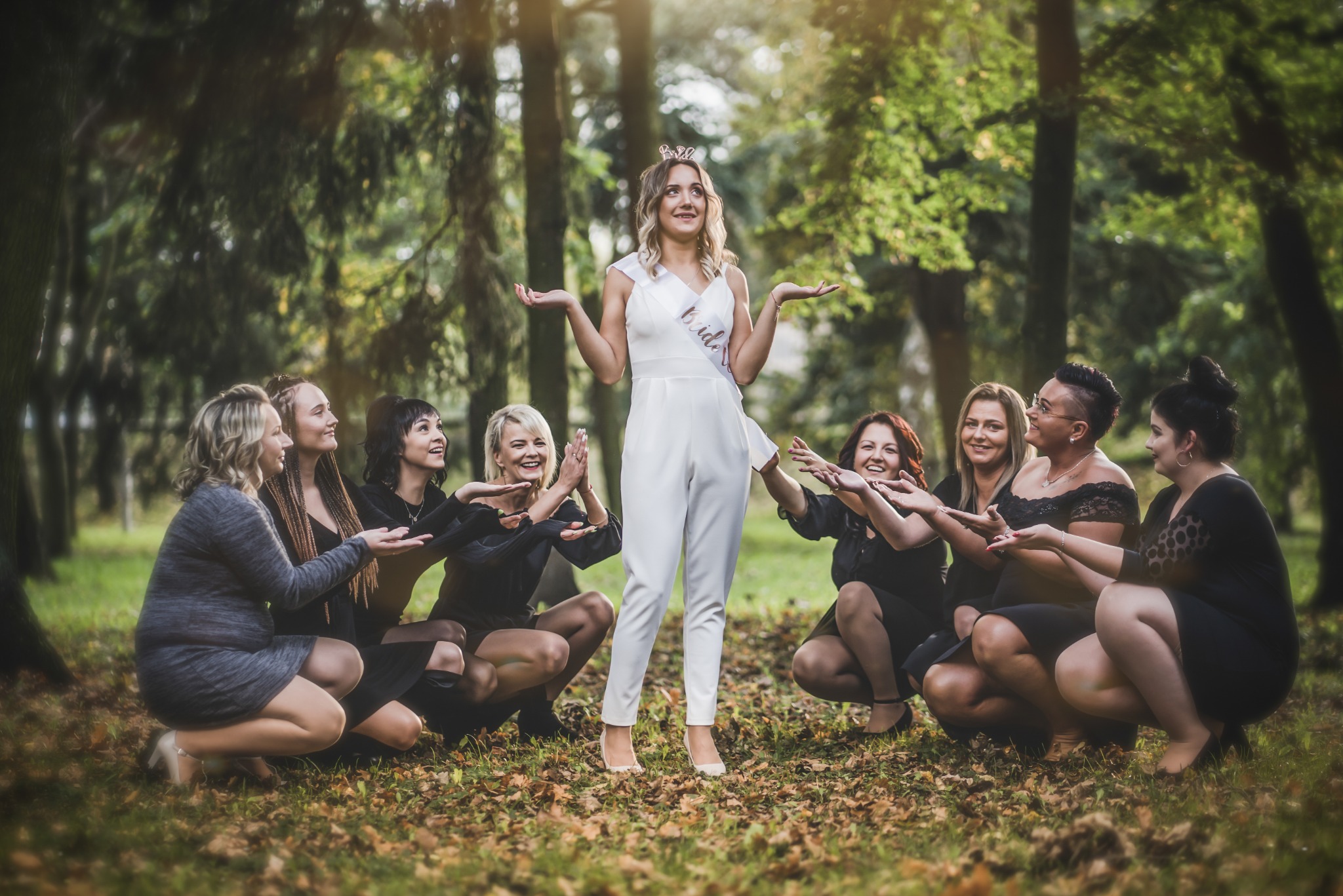 Panna młoda w białym kombinezonie i tiarze, otoczona przez druhny w czarnych sukienkach, pozuje na tle jesiennego lasu.