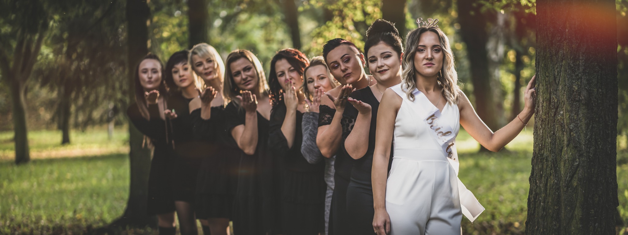 Panna młoda w białym kombinezonie z szarfą 'Bride to be' i koroną na głowie, pozuje z druhnami w czarnych sukienkach, wysyłając buziaki w plenerze.