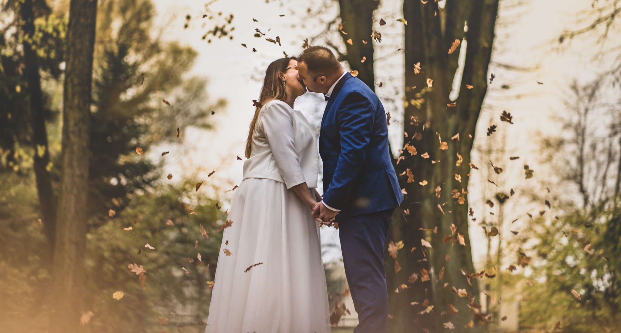 Pocałunek pary młodej w jesiennej scenerii, opadające liście tworzą romantyczną aurę, on w niebieskim garniturze, ona w białym komplecie, trzymają się za ręce.