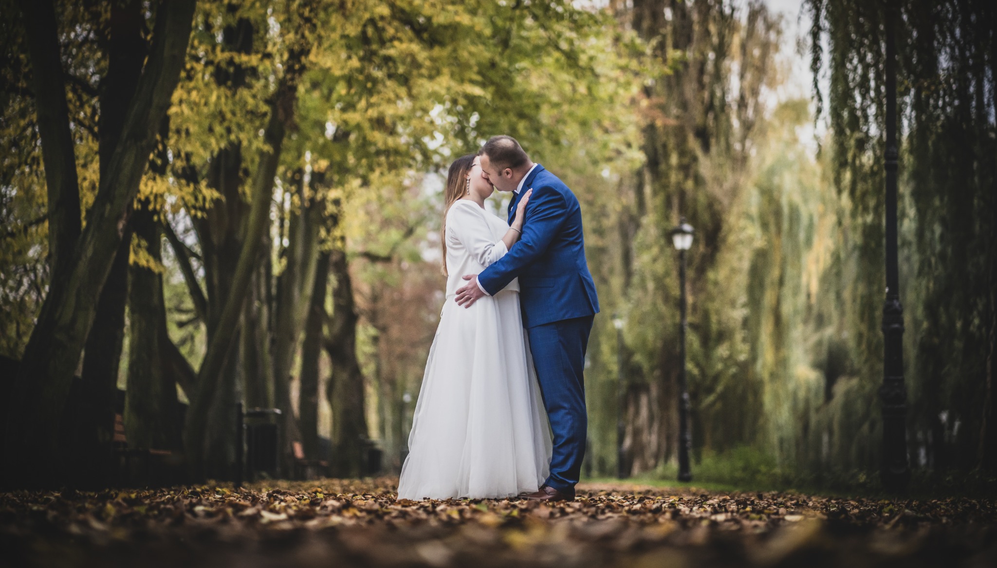 Pocałunek pary młodej w alei parkowej, on w niebieskim garniturze, ona w białej sukni i narzutce, jesienne liście na ziemi, w tle drzewa i latarnie.