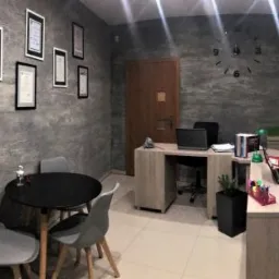 Wnętrze biura z szarymi ścianami o fakturze betonu, czarnym okrągłym stolikiem z szarymi krzesłami, biurkiem z laptopem i dokumentami oraz zegarem ściennym bez cyfr.