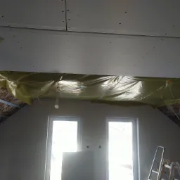 Poddasze w trakcie adaptacji: widoczna izolacja z wełny mineralnej, zabezpieczona folią, częściowo zakryta płytami kartonowo-gipsowymi, metalowa konstrukcja nośna, dwie okna dachowe i drabina...