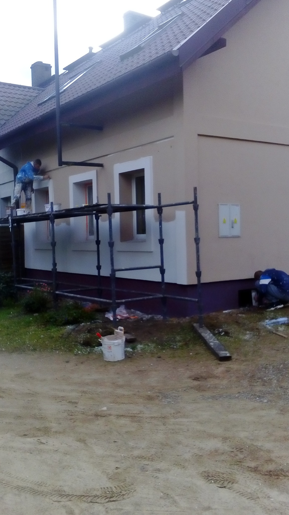 Dwóch pracowników na zewnątrz budynku, jeden na rusztowaniu maluje elewację okien, drugi przy ziemi, widoczny kubeł z farbą i narzędzia.