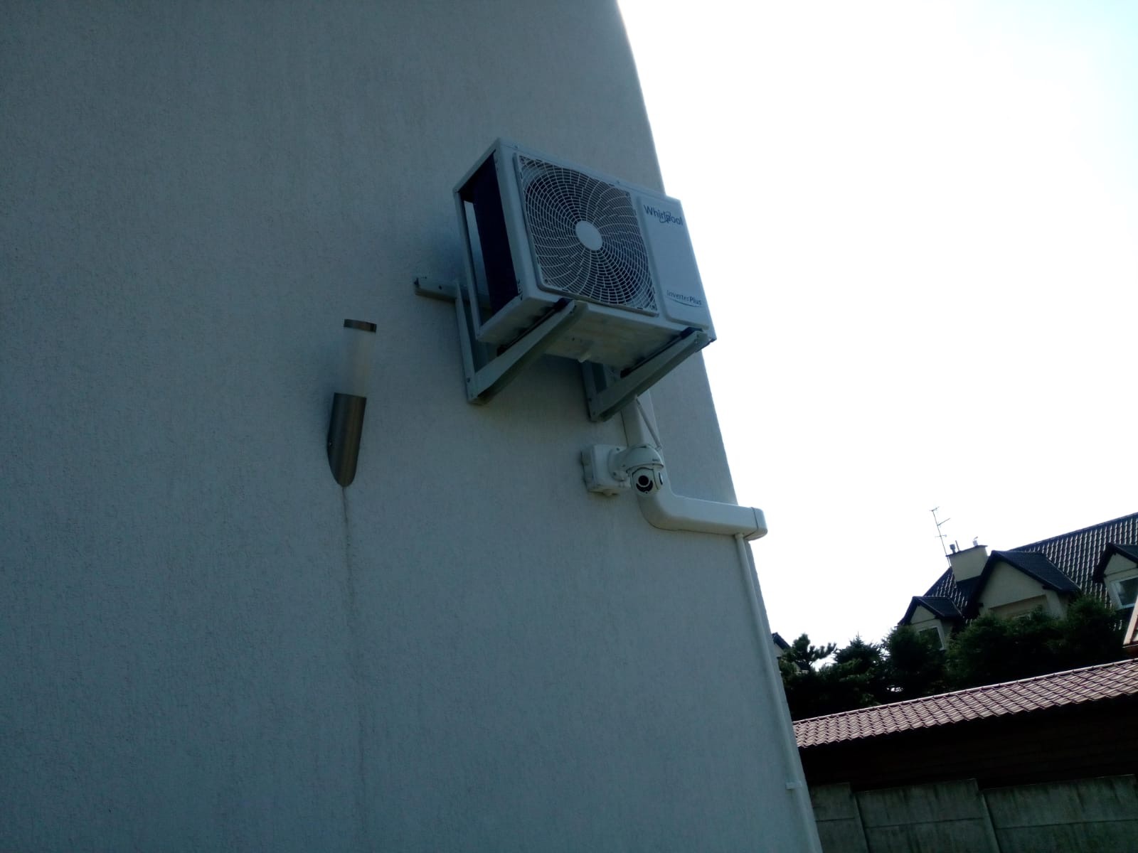 Biała jednostka zewnętrzna klimatyzatora Whirlpool zamontowana na ścianie budynku mieszkalnego, widoczna instalacja odprowadzająca skropliny i lampa ścienna.
