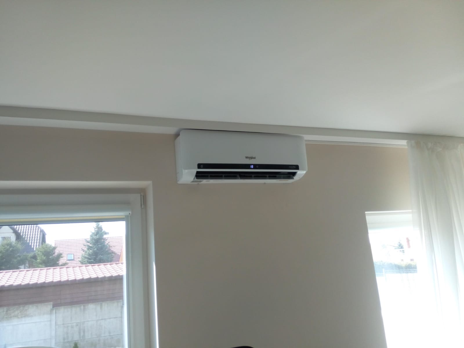 Biała klimatyzacja marki Whirlpool zamontowana na ścianie w jasnym pokoju, widoczne okno z widokiem na domy i drzewa, oraz biała zasłona.