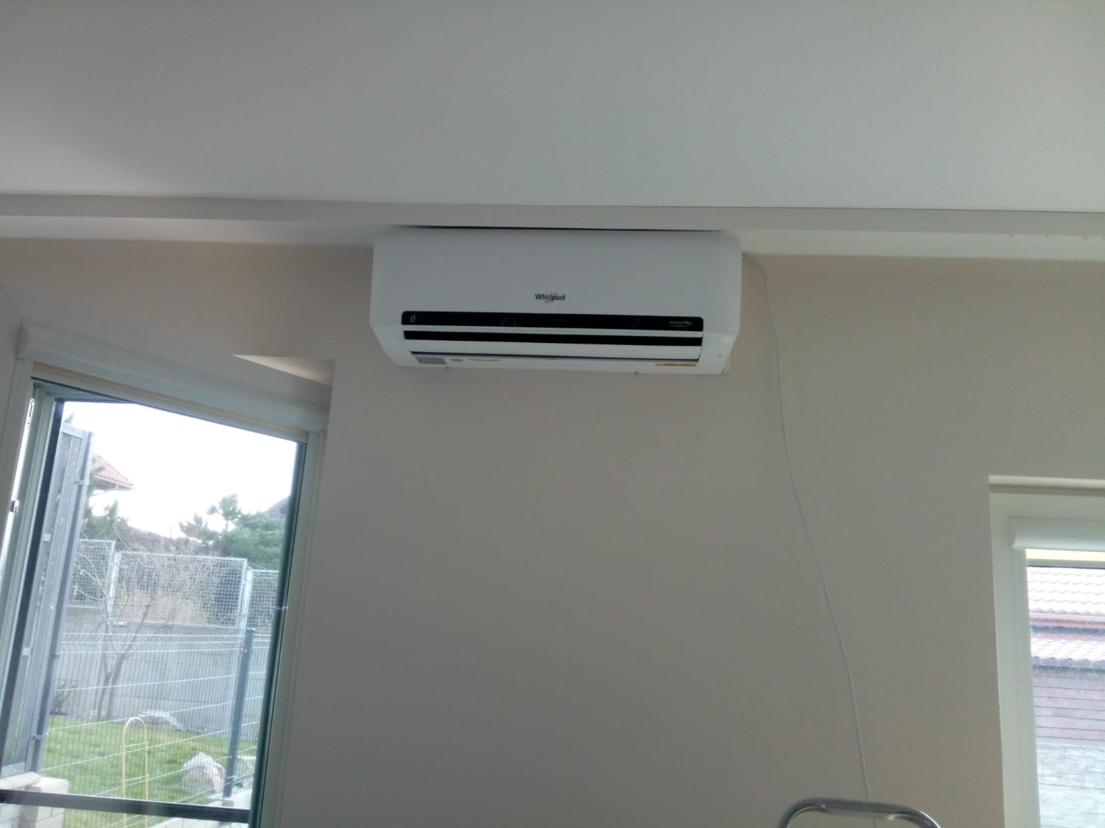 Biały klimatyzator Whirlpool zamontowany na ścianie w jasnym pomieszczeniu, widoczne okno z widokiem na ogród.