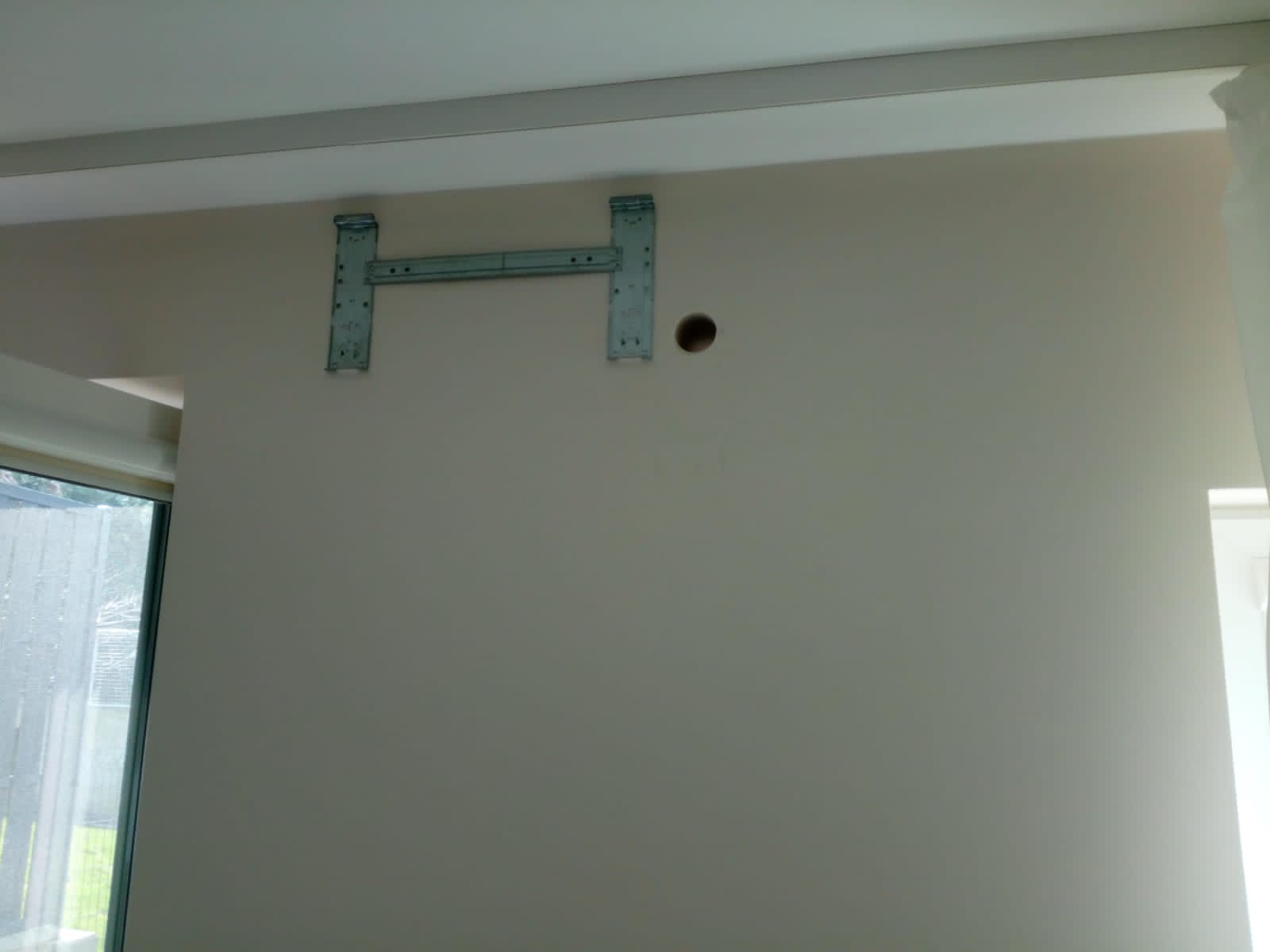 Metalowa konstrukcja wspornika klimatyzatora przymocowana do jasnej ściany wewnątrz pomieszczenia, widoczny otwór w ścianie obok wspornika.