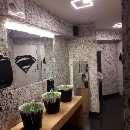 Nietypowa łazienka publiczna z trzema umywalkami w kształcie czarnych wiader z zieloną 'farbą' spływającą po brzegach, wzorzyste ściany imitujące marmur, lustro z namalowanym symbolem Supermana...