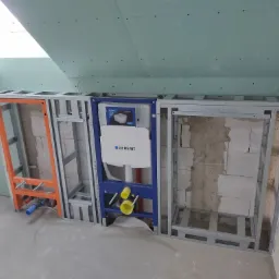 Stelaż instalacyjny podtynkowy Geberit Duofix zabudowany metalową konstrukcją, przygotowany do obłożenia płytami. Widoczne przyłącza wodne i kanalizacyjne, oraz fragment sufitu z zamontowanymi...