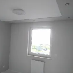 Jasne, pomalowane na szaro wnętrze pokoju z oknem, białym grzejnikiem pod parapetem i zamontowanym oświetleniem sufitowym.