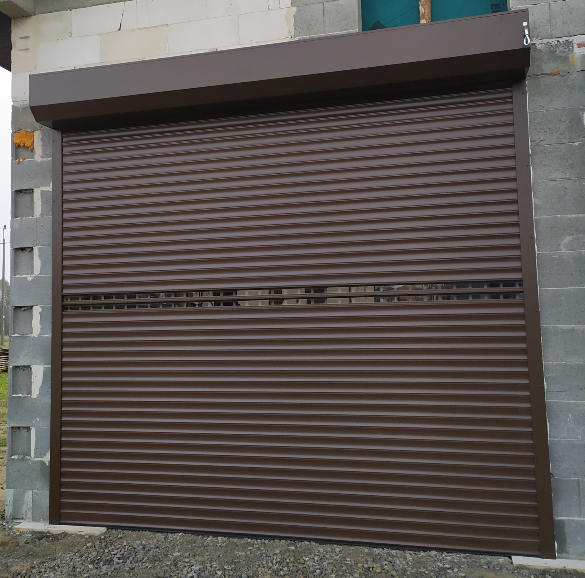 Brązowa roleta zewnętrzna częściowo opuszczona, widoczne luki między lamelami, zamontowana na surowym murze z betonu komórkowego.