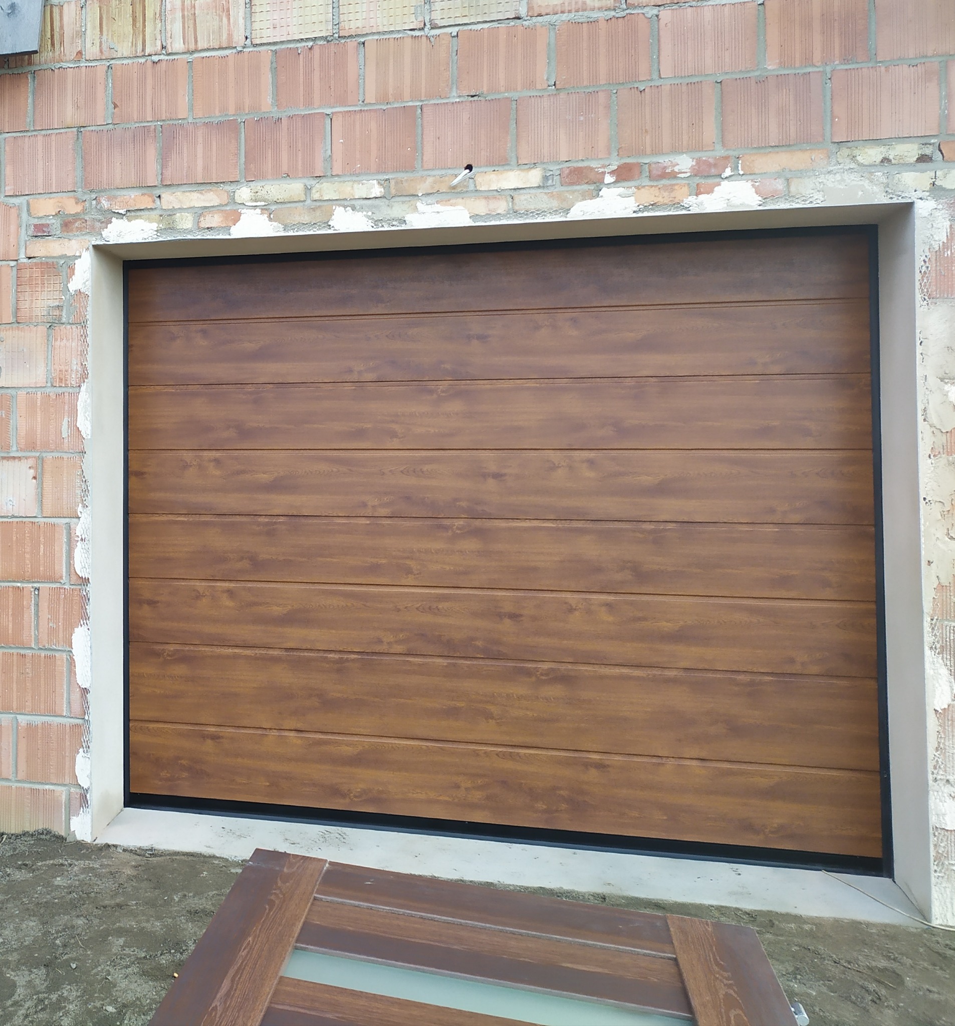 Brązowa segmentowa brama garażowa z efektem drewna, zamontowana w surowym murze z cegieł, fragment skrzydła bramy leżący na ziemi przed wjazdem.