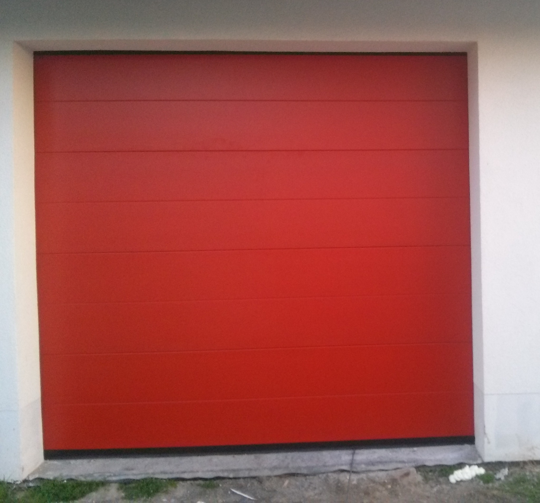 Czerwona, segmentowa brama garażowa z poziomymi panelami, widoczna z bliskiej odległości, zamontowana w białej ścianie budynku.