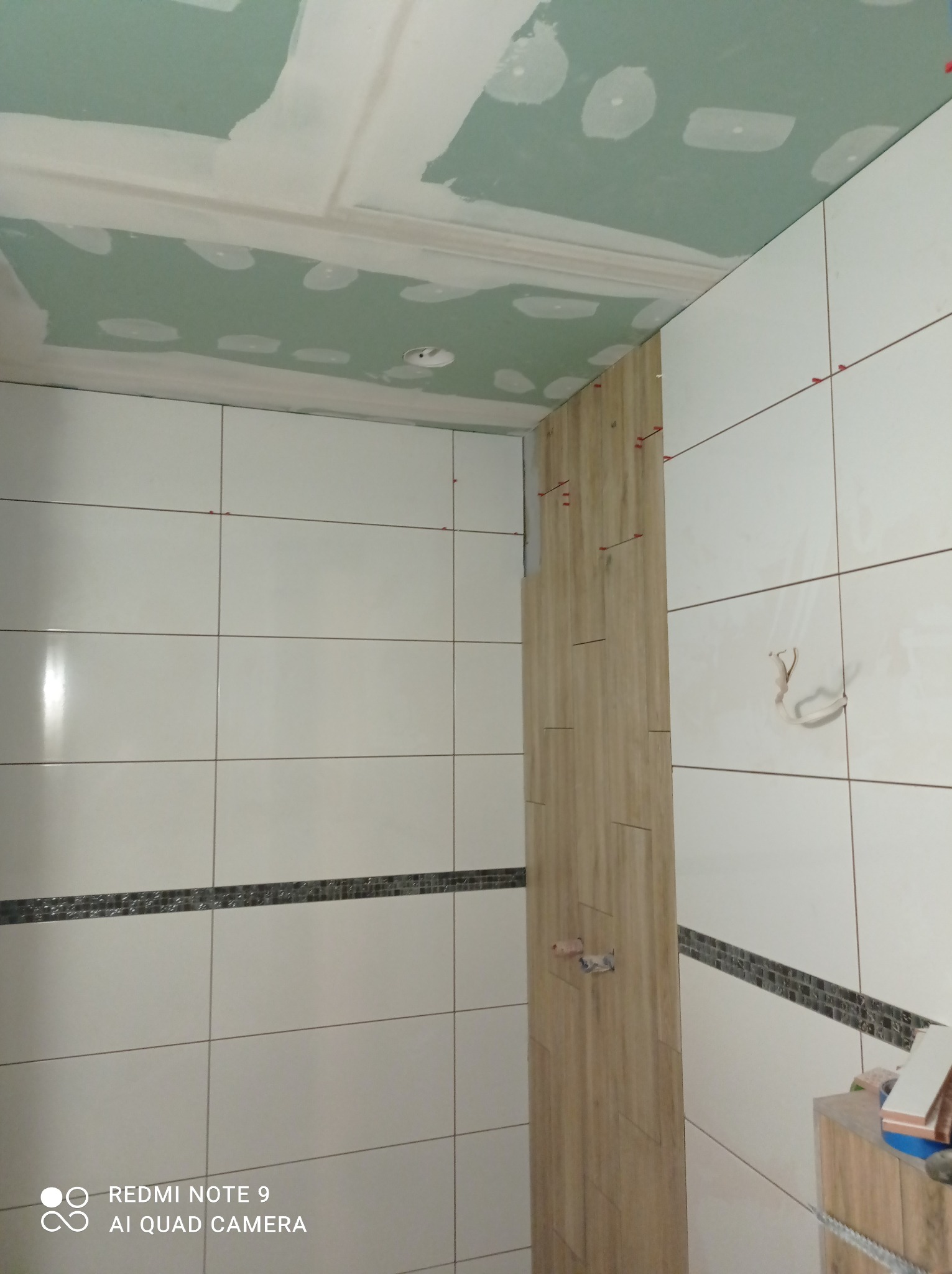 Wnętrze łazienki w trakcie wykańczania: białe płytki ceramiczne z dekoracyjnym pasem, fragment ściany z drewnopodobnymi płytkami, sufit z płyt kartonowo-gipsowych przed szpachlowaniem