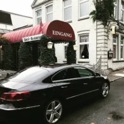 Czarny samochód zaparkowany przed wejściem do hotelu-restauracji z czerwonym baldachimem i napisem 'Eingang'.