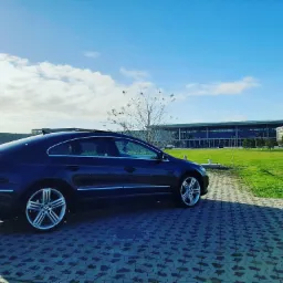 Granatowy samoch&oacute;d Volkswagen CC z otwartym szyberdachem, zaparkowany na brukowanej ścieżce z widokiem na nowoczesny budynek w tle, w słoneczny dzień.