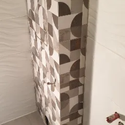 Obudowa pionu kanalizacyjnego wykończona płytkami z geometrycznym wzorem w odcieniach szarości i brązu, dopasowanymi do płytek na podłodze i ścianach łazienki, z widocznym otworem rewizyjnym...