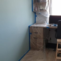 BCS Glazura i Wykończenia - Pokój przygotowany do malowania: ściana w kolorze błękitnym zabezpieczona taśmą malarską, meble i podłoga osłonięte folią ochronną.