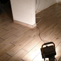 BCS Glazura i Wykończenia - Ułożona podłoga z płytek imitujących drewno w jasnym odcieniu, widoczna lampa budowlana oświetlająca fragment posadzki, ściany przygotowane do malowania.