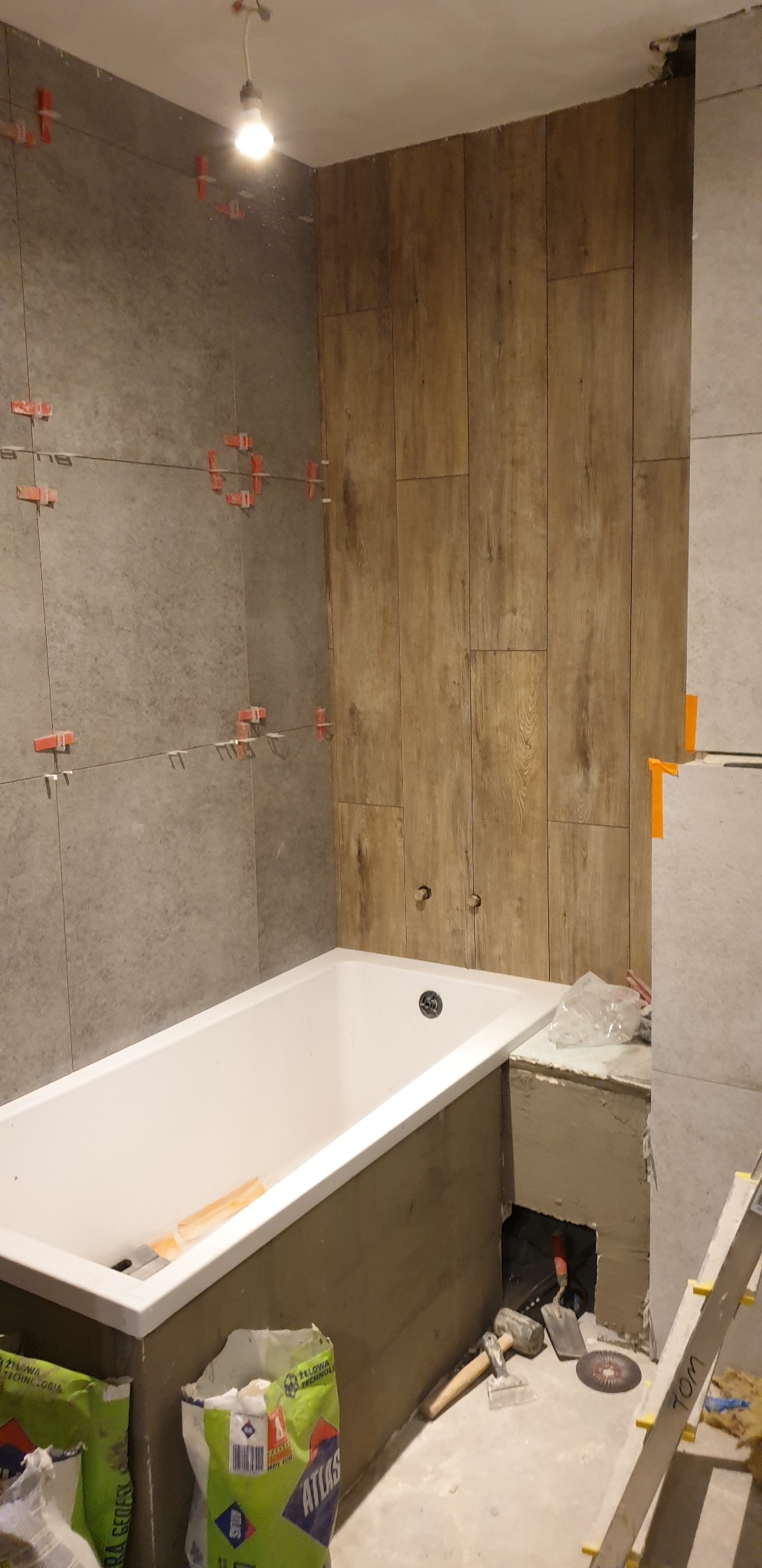 Montaż płytek imitujących drewno na ścianie w łazience, widoczna wanna, krzyżyki dystansowe, worki z klejem Atlas i narzędzia glazurnicze.