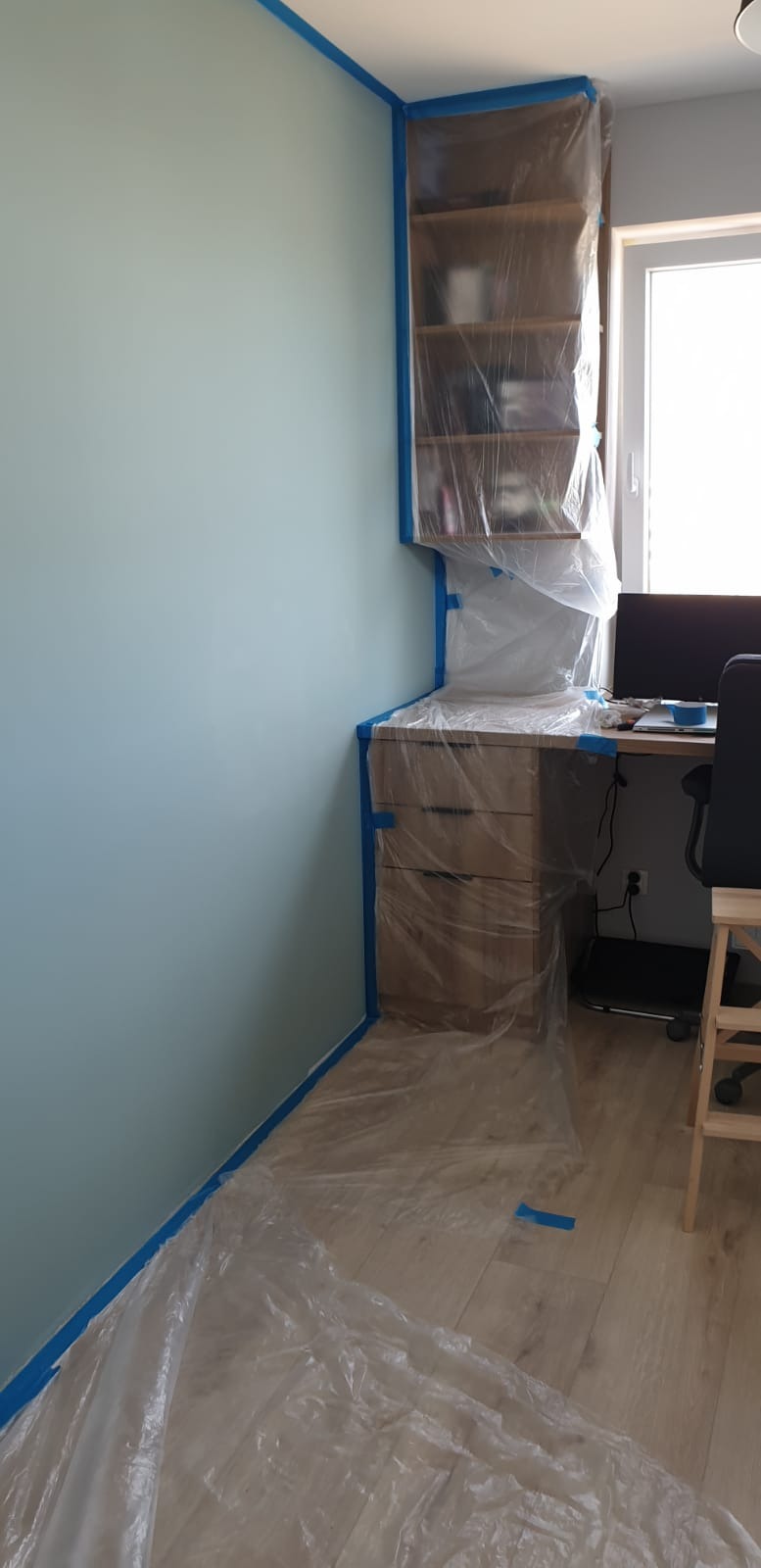 Pokój przygotowany do malowania: ściana w kolorze błękitnym zabezpieczona taśmą malarską, meble i podłoga osłonięte folią ochronną.
