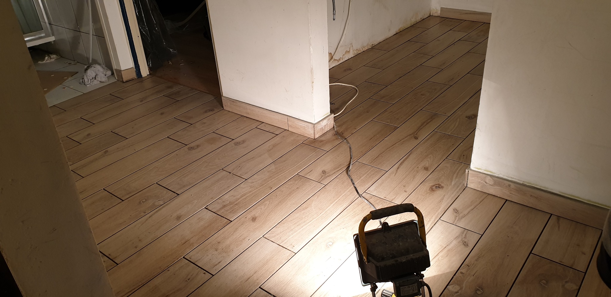 Ułożona podłoga z płytek imitujących drewno w jasnym odcieniu, widoczna lampa budowlana oświetlająca fragment posadzki, ściany przygotowane do malowania.