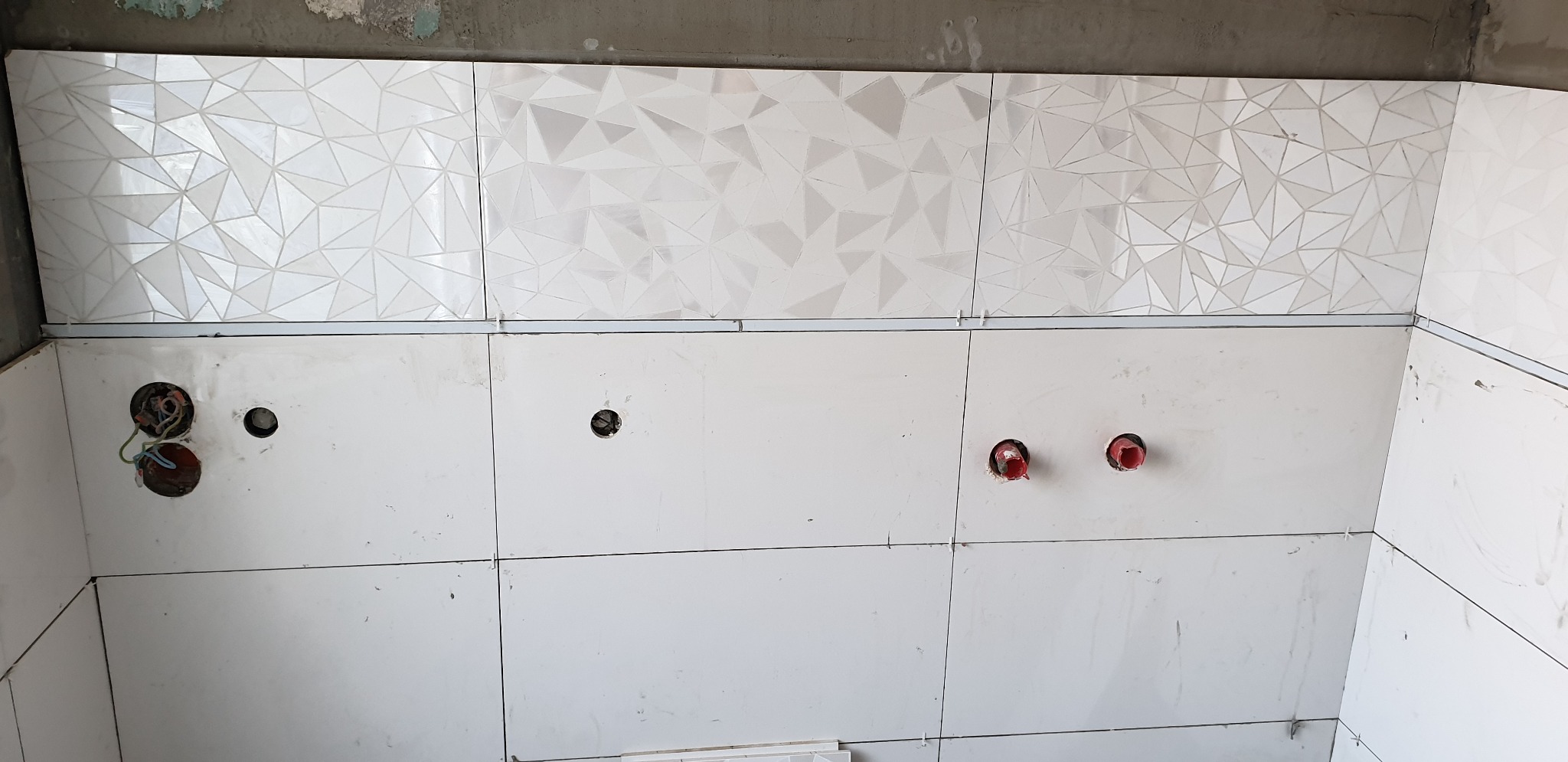 Ściana w trakcie wykańczania z białymi, błyszczącymi płytkami: górny rząd z geometrycznym wzorem, dolny rząd gładki, widoczne otwory na instalacje.