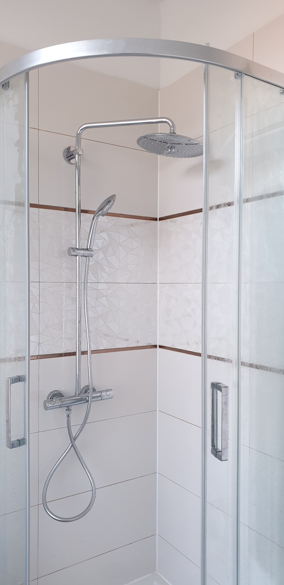 Kabina prysznicowa z chromowaną armaturą i białymi płytkami z geometrycznym wzorem oraz brązowym paskiem dekoracyjnym.