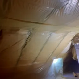 Poddasze zabezpieczone folią przed pracami ociepleniowymi, widoczne fragmenty konstrukcji dachu i materiały izolacyjne w rolkach.