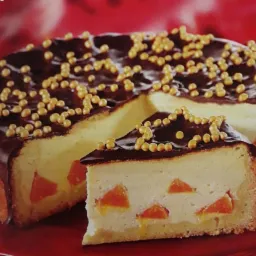 Tort serowy z polewą czekoladową i kandyzowanymi morelami, udekorowany złotymi kuleczkami, na czerwonym talerzu.