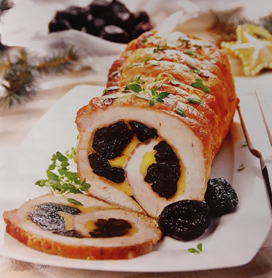 Pieczony schab rolowany z suszonymi śliwkami i serem, udekorowany świeżym tymiankiem na białym talerzu, obok leżą śliwki, w tle gałązka świerku i gwiazdka.