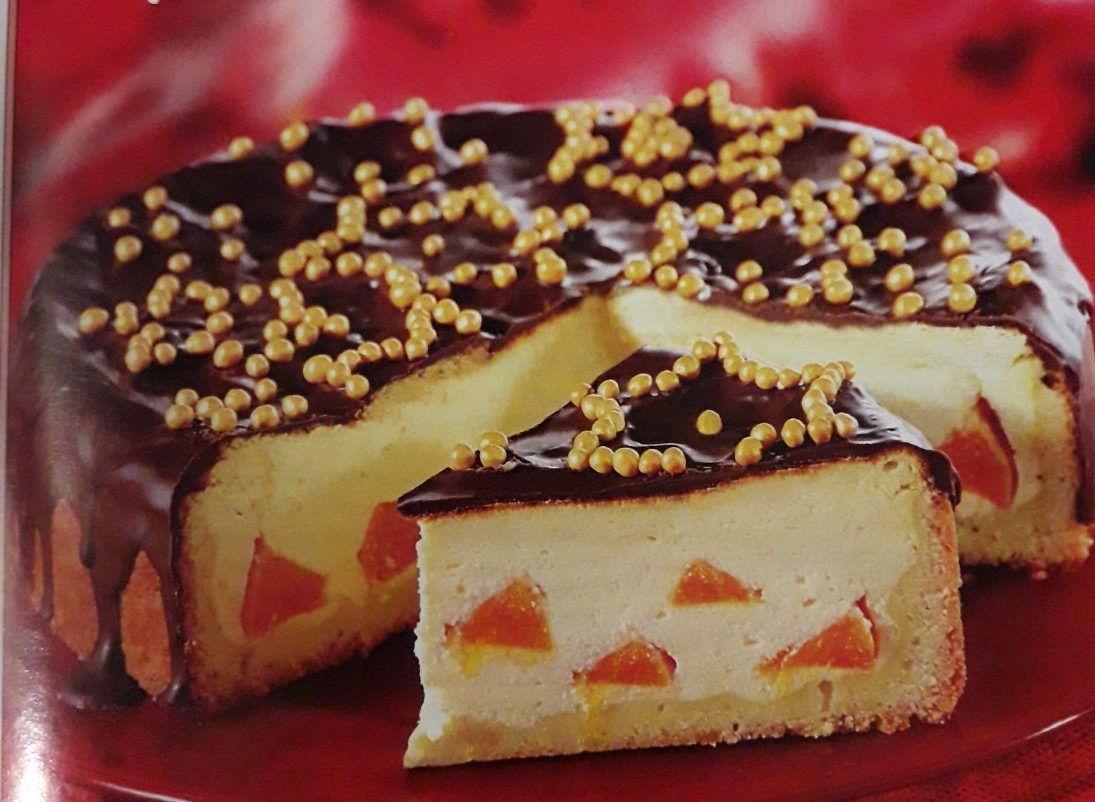 Tort serowy z polewą czekoladową i kandyzowanymi morelami, udekorowany złotymi kuleczkami, na czerwonym talerzu.