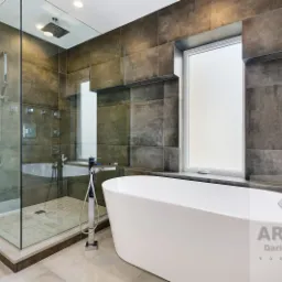 Nowoczesna łazienka z kabiną prysznicową typu walk-in z deszczownicą i panelami natryskowymi oraz wolnostojącą białą wanną, wykończona ciemnymi płytkami imitującymi beton i jasnymi płytkami...