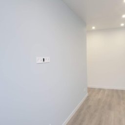 Ryskin .sp.z.o.o - Świeżo pomalowane, jasnoniebieskie ściany w pustym pokoju z drewnopodobną podłogą, widoczne gniazdka elektryczne w białej ramce.