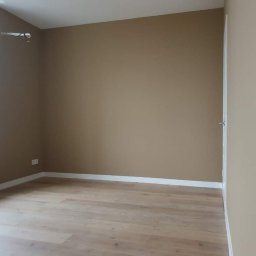 Ryskin .sp.z.o.o - Wnętrze pomalowanego na brązowo pokoju w Gdańsku z drewnopodobną podłogą i białymi listwami przypodłogowymi, widoczny grzejnik pod oknem.