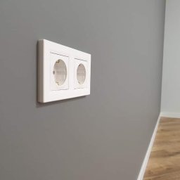 Ryskin .sp.z.o.o - Dwa białe gniazdka elektryczne zamontowane na świeżo pomalowanej szarej ścianie, widoczny fragment drewnianej podłogi przy białej listwie przypodłogowej.