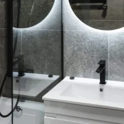 Nowoczesna łazienka z umywalką nablatową, czarną armaturą i podświetlanym lustrem w kształcie półksiężyca na tle płytek imitujących kamień.