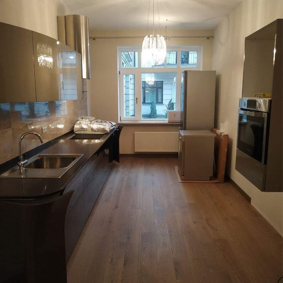 Ciemna, nowoczesna zabudowa kuchenna z błyszczącymi frontami, stalowym okapem i drewnianą podłogą w trakcie montażu, z widocznymi kartonami i zabezpieczeniami na blacie.