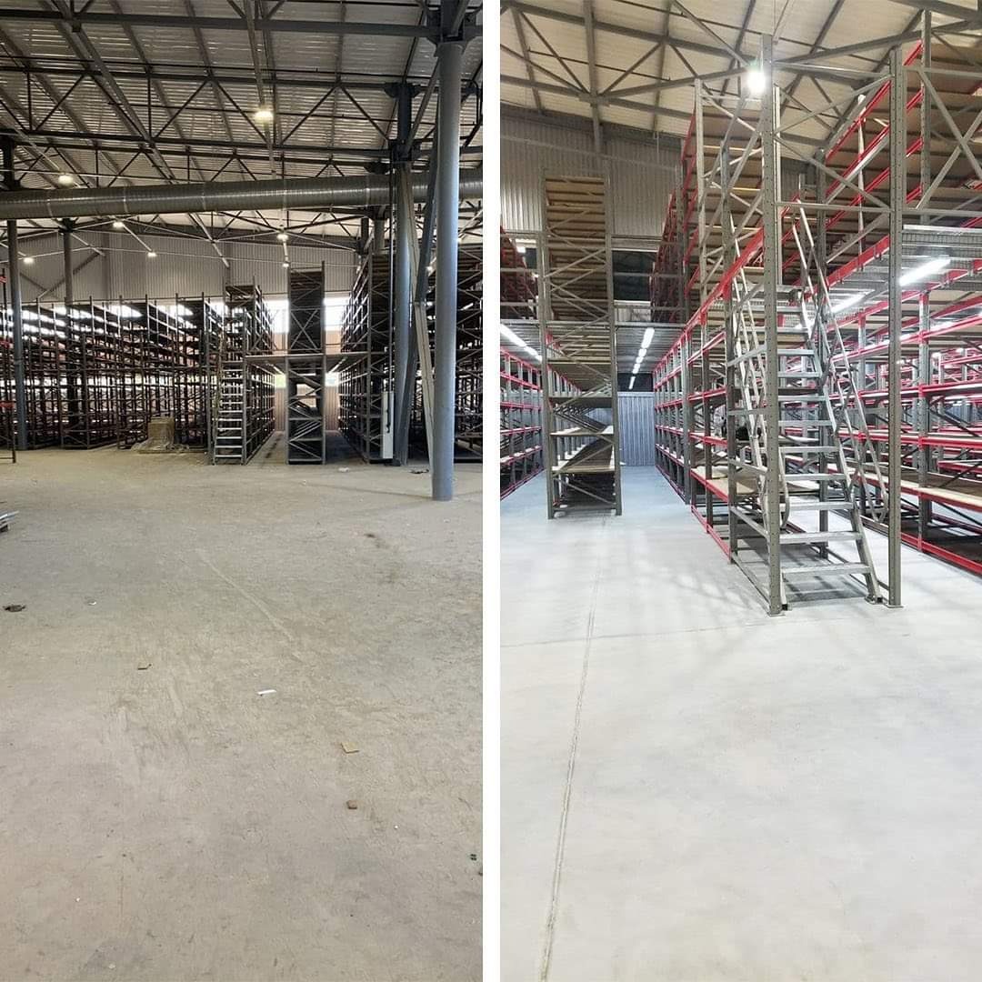 Rozległa hala magazynowa z metalowymi regałami, podzielona na dwie części: starsza z szarymi regałami i nowsza z czerwonymi akcentami, oświetlona lampami sufitowymi.