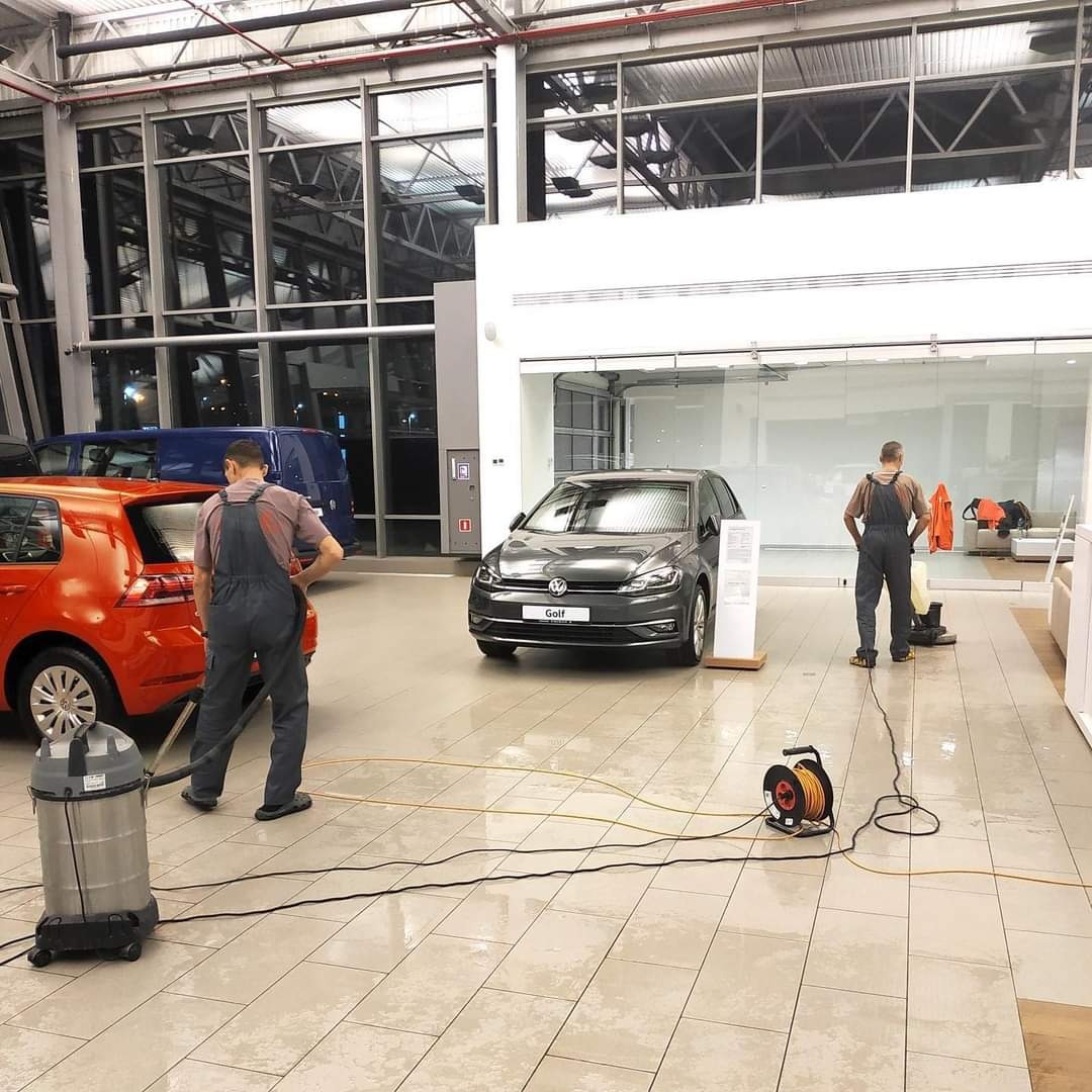 Dwóch mężczyzn w kombinezonach czyści podłogę w salonie samochodowym, widoczne dwa samochody osobowe, odkurzacz przemysłowy i zwinięty przedłużacz.