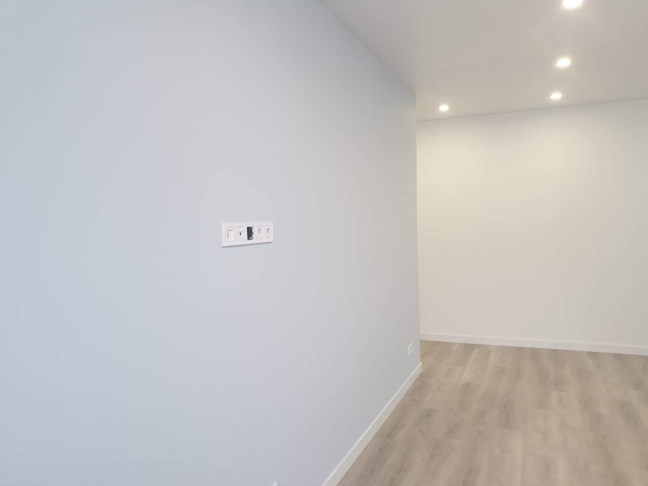 Świeżo pomalowane, jasnoniebieskie ściany w pustym pokoju z drewnopodobną podłogą, widoczne gniazdka elektryczne w białej ramce.