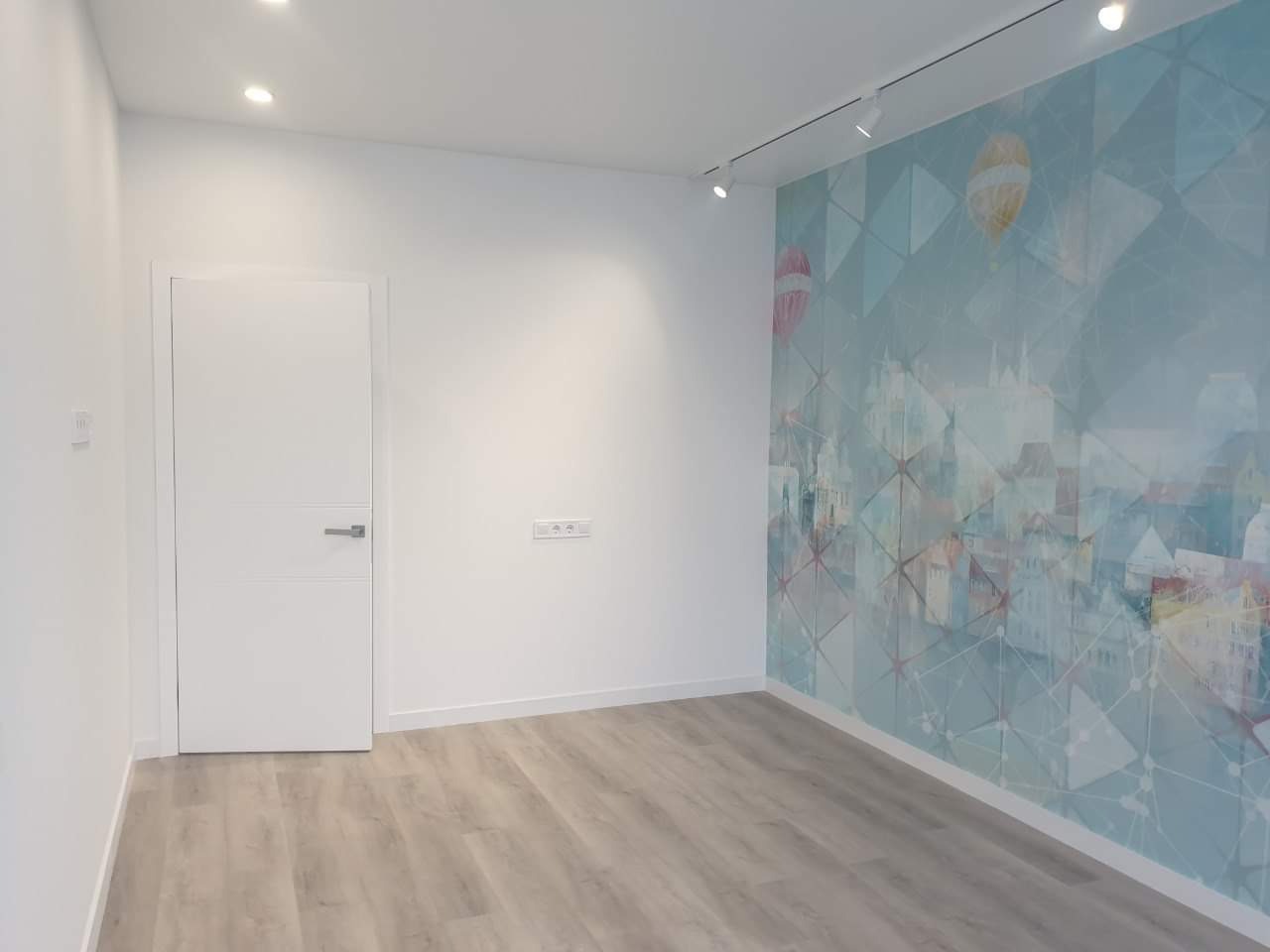 Jasne wnętrze pokoju z drewnianą podłogą i ścianą ozdobioną fototapetą z motywem miasta i balonów, oświetlone punktowymi lampami sufitowymi.