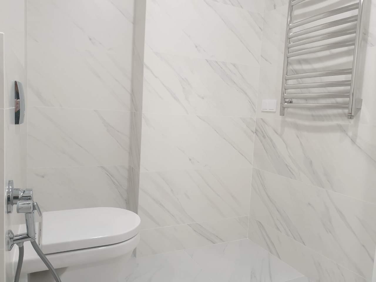 Biała łazienka z marmurowymi płytkami, podwieszaną toaletą i grzejnikiem drabinkowym.