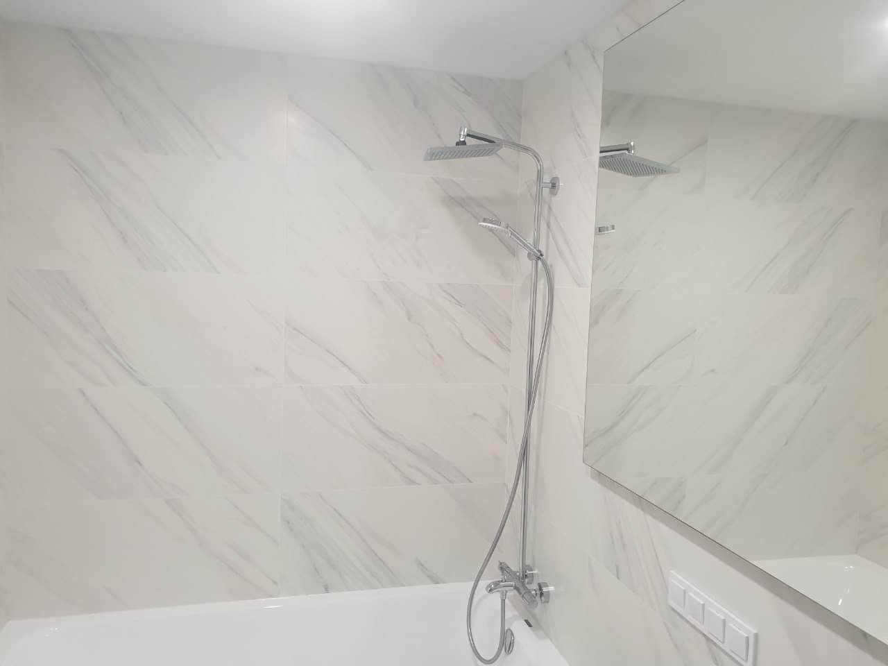 Biała łazienka z wanną, prysznicem deszczownicą i płytkami imitującymi marmur, odbicie w lustrze. Minimalistyczny design.