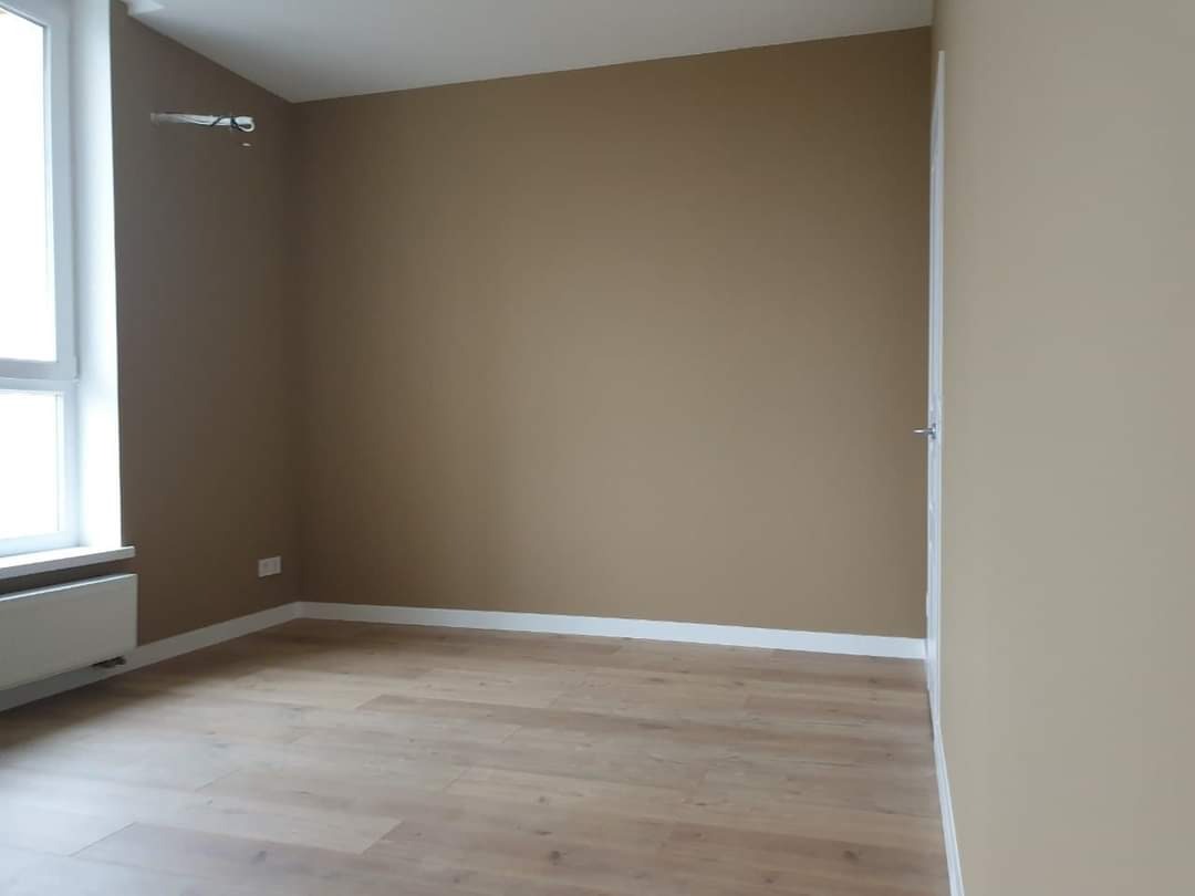 Wnętrze pomalowanego na brązowo pokoju w Gdańsku z drewnopodobną podłogą i białymi listwami przypodłogowymi, widoczny grzejnik pod oknem.