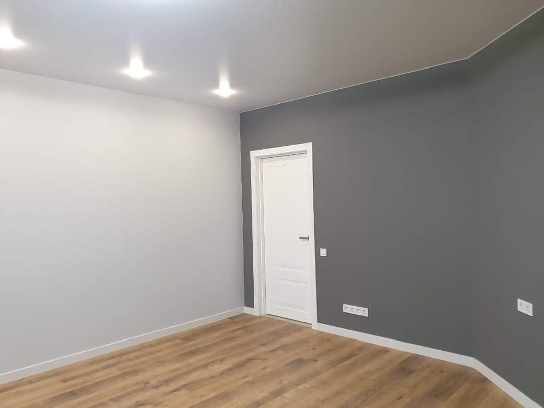 Pokój po remoncie z dwiema ścianami pomalowanymi na różne kolory, jedna szara, druga biała, drewniana podłoga, białe drzwi i listwy przypodłogowe, trzy punktowe lampy na suficie.