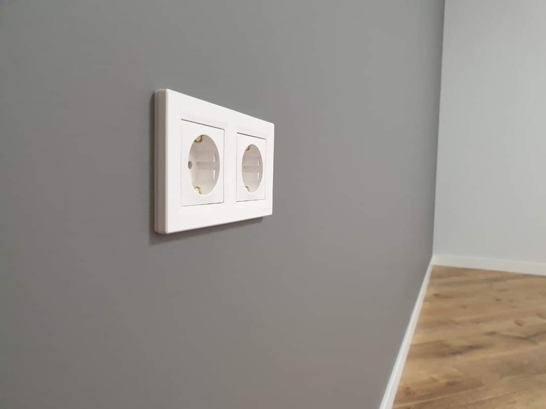 Dwa białe gniazdka elektryczne zamontowane na świeżo pomalowanej szarej ścianie, widoczny fragment drewnianej podłogi przy białej listwie przypodłogowej.