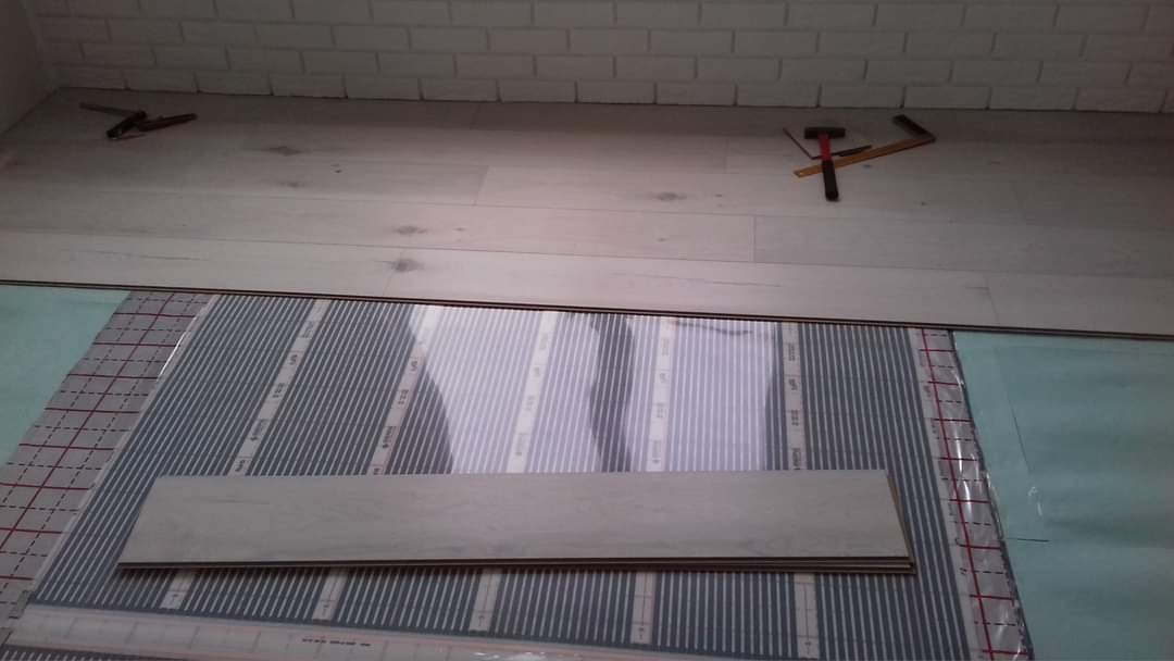 Podkład pod panele podłogowe z systemem ogrzewania podłogowego, widoczne narzędzia i fragment ściany wykończonej cegłą.